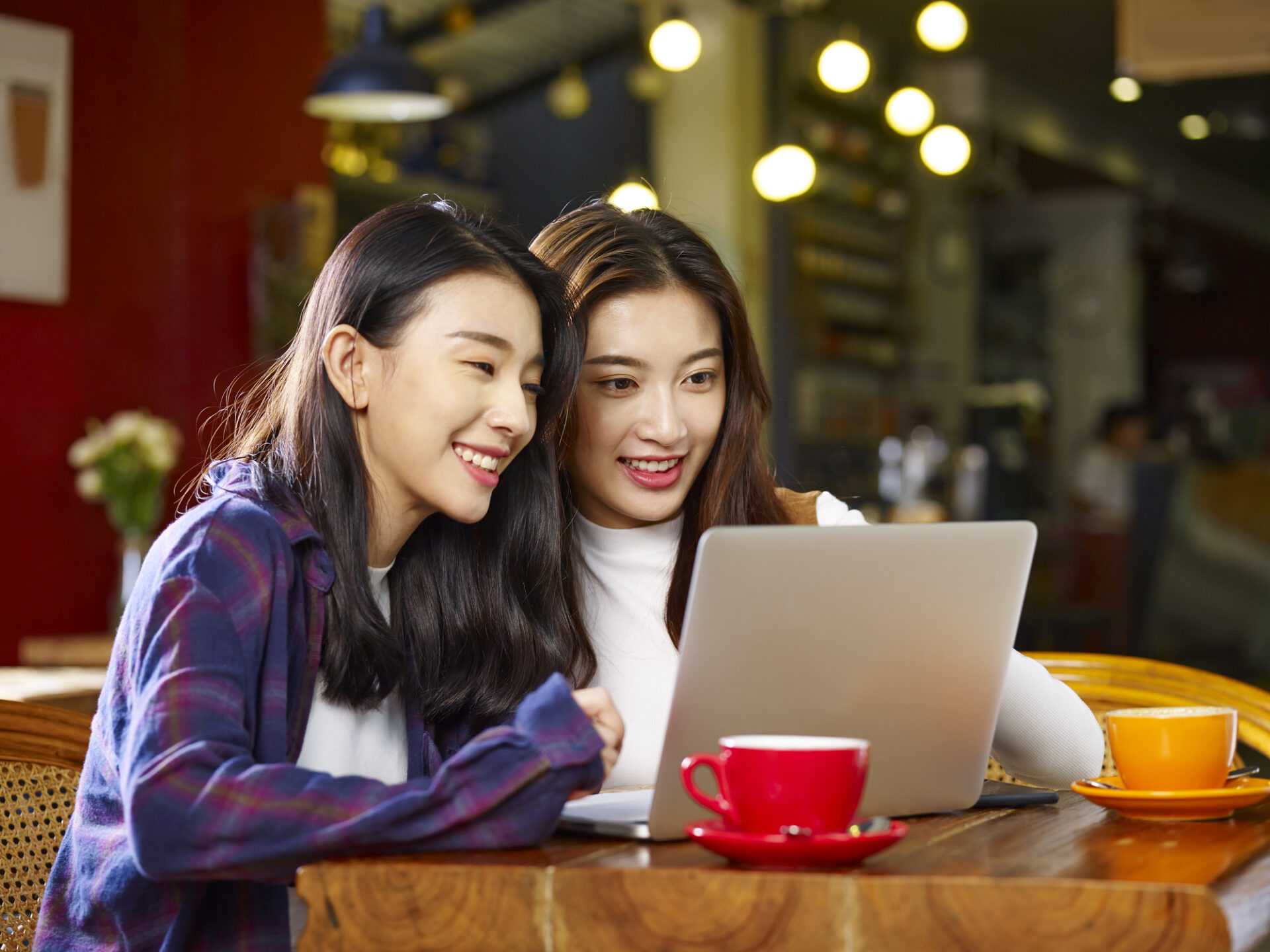 two happy young asian women sitting in coffee shop looking at laptop computer together.